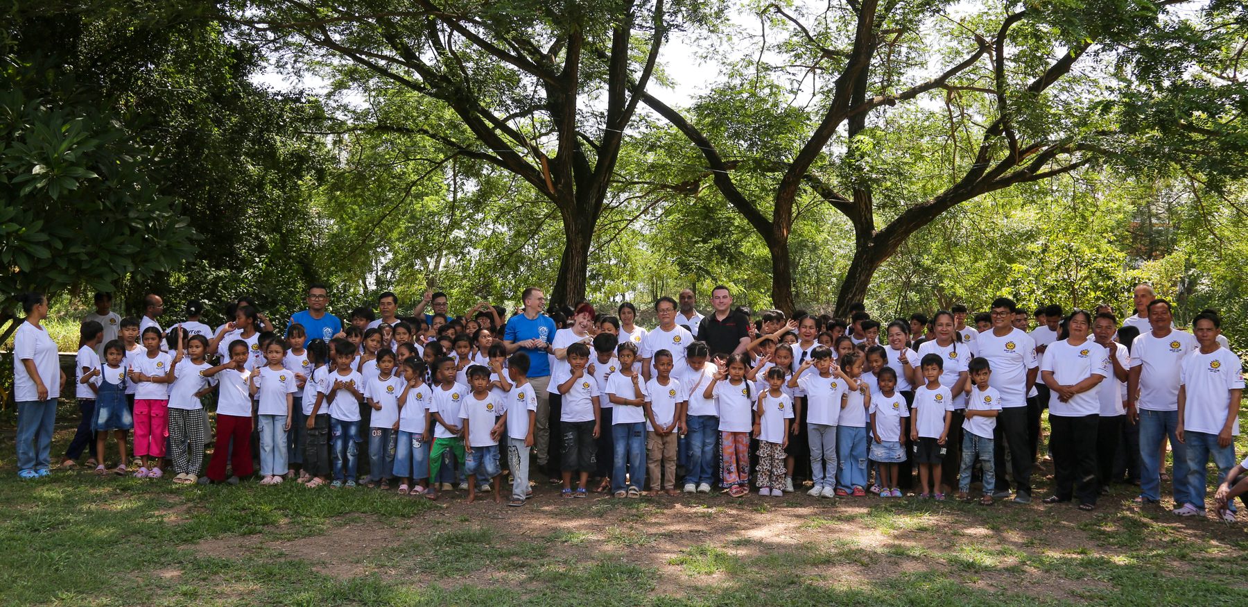 A very large group of children and staff in white shirts standing together under big trees at Sunrise Cambodia