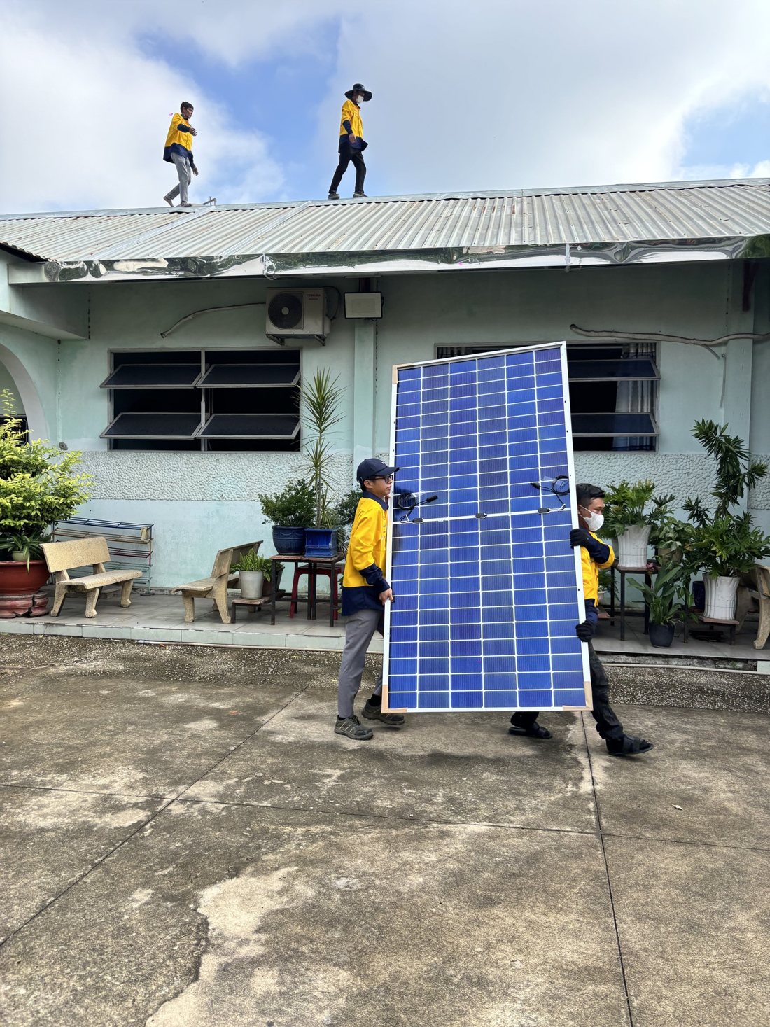 Two installers carrying a solar panel across a courtyard while a colleague walks the roof above