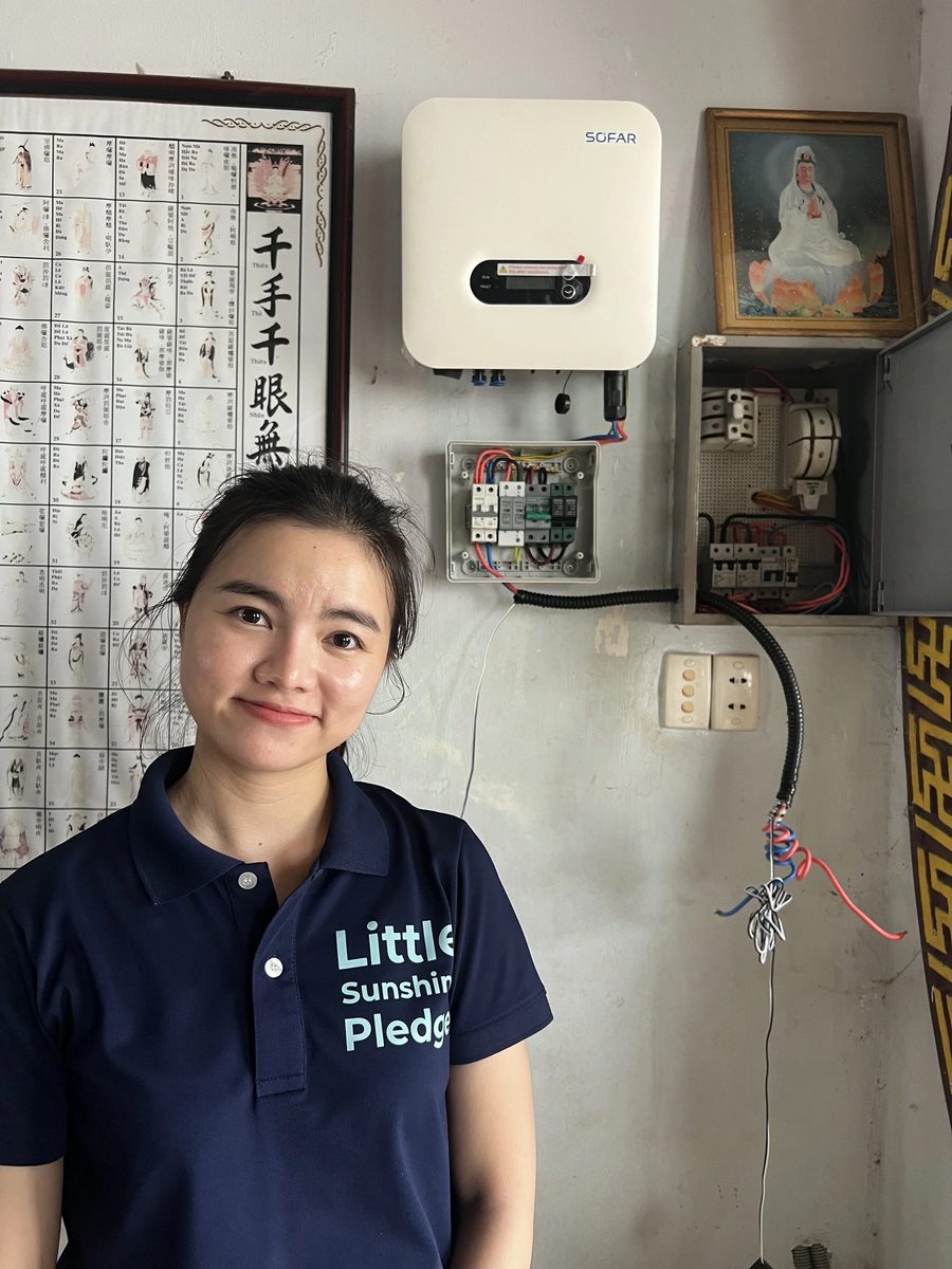 A team member in a navy charity shirt beside a newly installed solar inverter on a wall