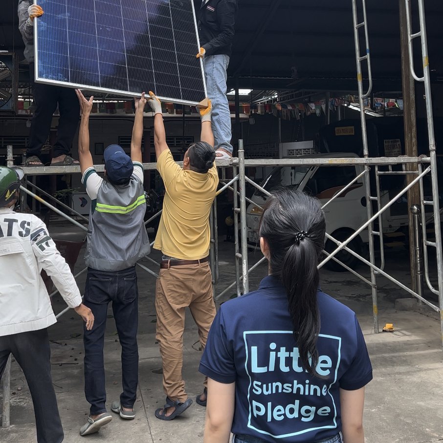 Installers lifting a solar panel up to a scaffold platform while a volunteer in a Little Sunshine Pledge shirt watches from behind