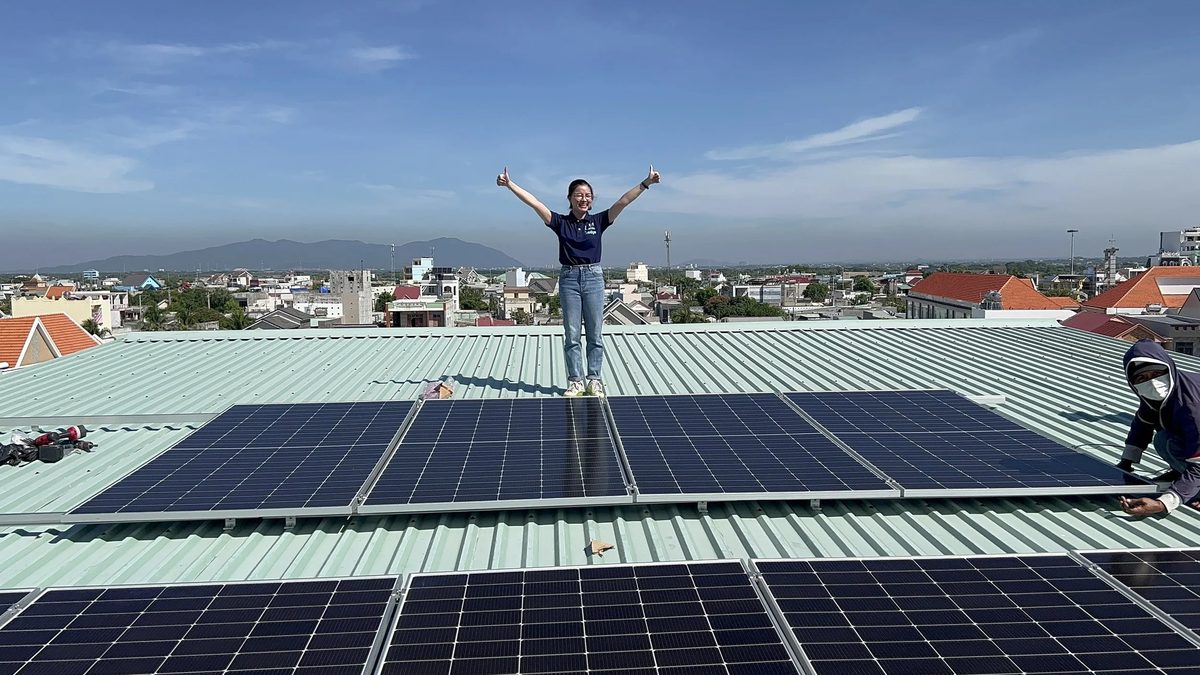 A team member in a navy charity shirt standing among solar panels on a roof with arms stretched out under a blue sky