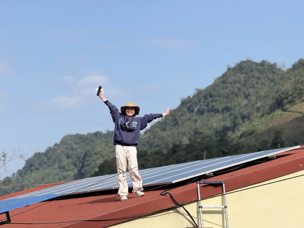 A person in a sun hat on a red roof with solar panels, green mountains behind
