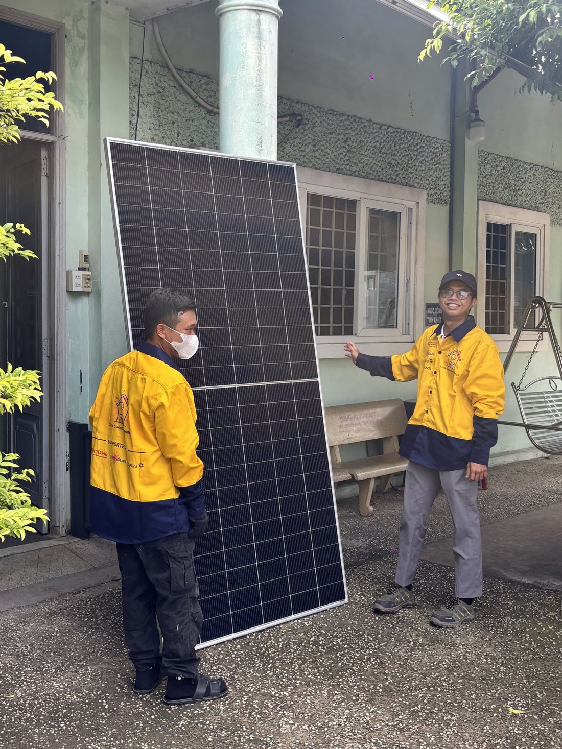 Two installers in yellow Little Sunshine Pledge work shirts standing beside a solar panel, one smiling